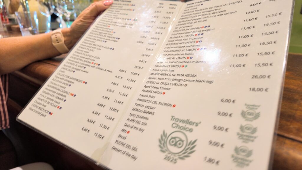 AbraCasaBra.se En person håller i en meny på en restaurang, med flera mat- och dryckesalternativ listade på spanska. På menyn finns priser och vissa rätter markerade med ikoner, samt ett Travellers' Choice 2023-märke på höger sida.