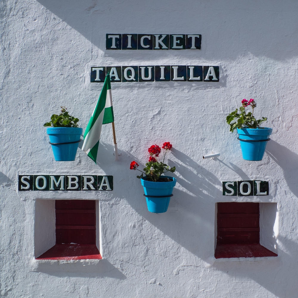 AbraCasaBra.se En vit vägg med två små fönster, tre blå blomkrukor med röda och gröna växter, en grönvit flagga och svarta skyltar med texten TICKET, TAQUILLA, SOMBRA och SOL.