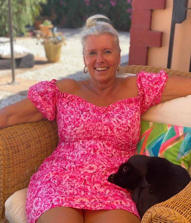 AbraCasaBra.se En leende kvinna i rosa blommig klänning sitter på en korgstol utomhus med en liten svart hund vilande i knät. Färgglada kuddar och krukväxter finns i bakgrunden.